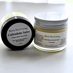 May include: Two glass jars of Savvy Eco Living Calendula Salve. The left jar has a black lid and a 1 oz label. The right jar has a white lid and a 2 oz label. Both labels list ingredients including plant-based oils, calendula herb, and beeswax.