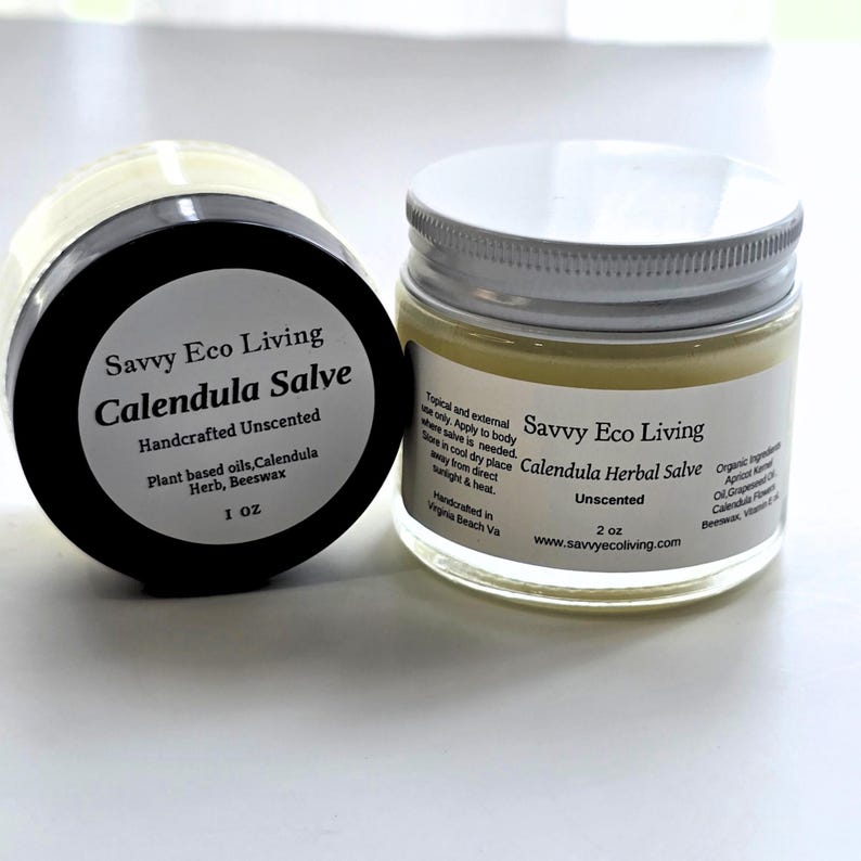 May include: Two glass jars of Savvy Eco Living Calendula Salve. The left jar has a black lid and a 1 oz label. The right jar has a white lid and a 2 oz label. Both labels list ingredients including plant-based oils, calendula herb, and beeswax.