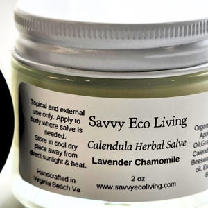 May include: A clear glass jar with a white screw-top lid containing a pale yellow herbal salve. The label reads Savvy Eco Living, Calendula Herbal Salve, Lavender Chamomile, 2 oz, and includes usage instructions and website information.