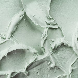 May include: A thick, pale sage green substance with a dense, creamy texture. The surface features prominent, sweeping ridges and peaks created by a spatula or tool, showing a matte finish with subtle variations in light and shadow across the swirls.