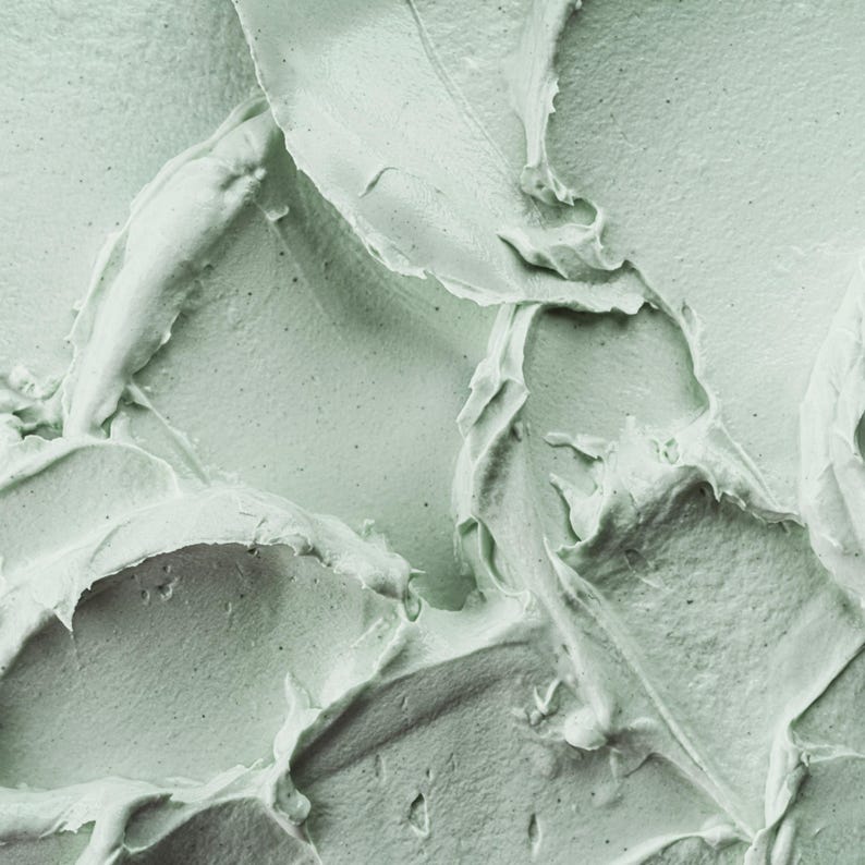 May include: A thick, pale sage green substance with a dense, creamy texture. The surface features prominent, sweeping ridges and peaks created by a spatula or tool, showing a matte finish with subtle variations in light and shadow across the swirls.