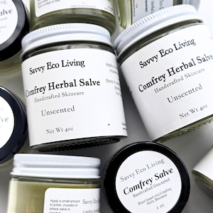 May include: Multiple glass jars of Savvy Eco Living Comfrey Herbal Salve with white screw-top lids. The labels feature black text on a white background, stating Handcrafted Skincare, Unscented, and Net Wt 4oz or 1oz, with visible ingredient lists.
