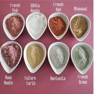 May include: Eight white teardrop-shaped bowls containing various colored cosmetic clays. The labels read: French Pink, White Kaolin, French Red, Rhassoul, Rose Kaolin, Fullers Earth, Bentonite, and French Green. The powders are fine and textured.