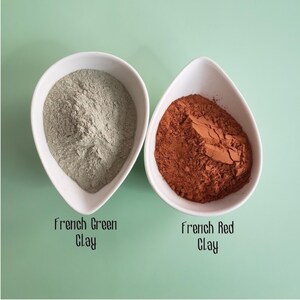 May include: Two white teardrop-shaped bowls containing fine powder. The left bowl holds light sage green powder labeled French Green Clay, and the right bowl holds deep terracotta red powder labeled French Red Clay, both on a light green surface.