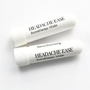 K&ouml;nnte beinhalten: Zwei wei&szlig;e Aromatherapie-Inhalatoren mit dem Text "HEADACHE EASE" und "Aromatherapy Inhaler" in Schwarz aufgedruckt. Ein Inhalator tr&auml;gt auch den Text "Savvy Eco Living". Die Inhalatoren haben eine zylindrische Form mit geriffelter Kappe.