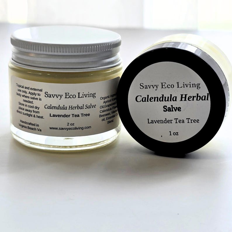 May include: Two clear glass jars of Savvy Eco Living Calendula Herbal Salve with Lavender Tea Tree. The left jar has a white lid and a 2 oz label with usage instructions. The right jar has a black lid and a 1 oz label showing the product name.