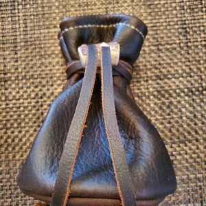 May include: A dark brown leather pouch with a drawstring closure. The pouch features a decorative antler clasp and is stitched with white thread. The leather has a textured appearance, and the drawstring is made of brown leather.