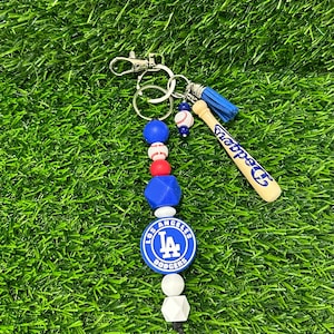 May include: A blue and white silicone beaded keychain with a baseball charm, a blue tassel, and a wooden baseball bat charm with the text "Choppers" on it. The keychain also features a blue and white silicone bead with the Los Angeles Dodgers logo.