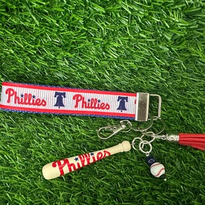 May include: A red, white, and blue keychain with a baseball bat charm, a baseball charm, and a tassel. The keychain has the words "Phillies" and a Liberty Bell graphic.