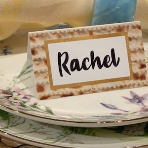 May include: A white place card with a gold border and a matzah texture background. The name "Rachel" is written in black cursive.