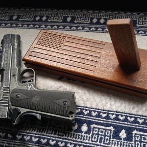 May include: A wooden gun display stand with an American flag design. The stand has a vertical support for the gun and a base with the flag design. A black handgun is lying on the stand.