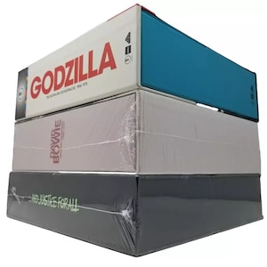 May include: Three stacked vinyl record boxes. The top box is teal blue with the text "GODZILLA THE SHOWA ERA SOUNDTRACKS 1954-1975". The middle box is light gray with the text "DAVID BOWIE" and "SOUNDTRACK (1983-2001)". The bottom box is dark gray with the text "...AND JUSTICE FOR ALL".