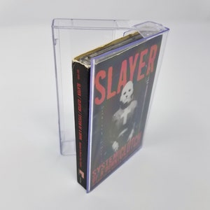 May include: A clear plastic cassette tape case with a black cassette tape inside. The cassette tape has a black and white photo of a person wearing a mask and a red and white label that says "SLAYER /  SYSTEM OF A DOWN" and "SYSTEMICLUTION OF A DOWN".