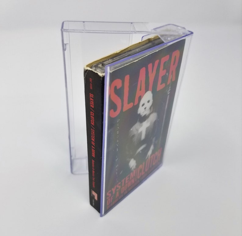May include: A clear plastic cassette tape case with a black cassette tape inside. The cassette tape has a black and white photo of a person wearing a mask and a red and white label that says "SLAYER /  SYSTEM OF A DOWN" and "SYSTEMICLUTION OF A DOWN".