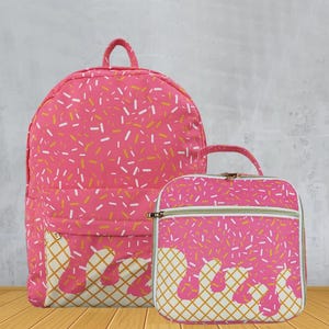 May include: Pink backpack and matching lunch bag set. Both items feature a pink background with white and yellow sprinkles. The bottom of the backpack and lunch bag have a waffle cone design with dripping ice cream.