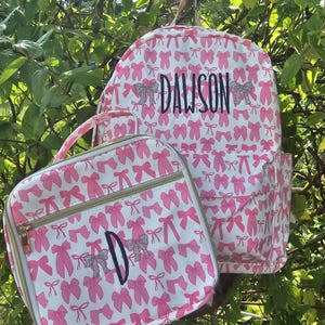 May include: A white backpack and lunch bag set with a pink bow pattern. The backpack has the name "DAWSON" in black script. The lunch bag has the letter "D" in black script. Both bags have a gold zipper.