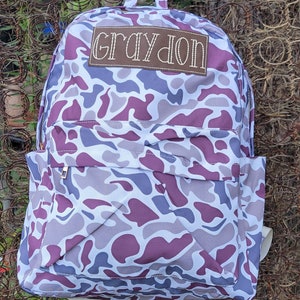 May include: A backpack with a camouflage pattern in shades of white, purple, and gray. The backpack has a front pocket with a zipper, side pockets, and adjustable straps. The name "GRAYSON" is embroidered on a brown patch on the front.