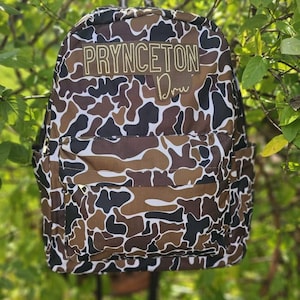 May include: A camouflage-patterned backpack with brown, black, and white shapes. The backpack has the text "PRYNCETON" in gold lettering across the top and "Dru" in cursive below. The backpack is hanging in front of green foliage.