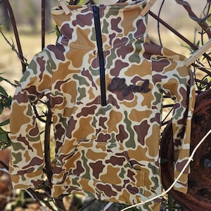 May include: A camouflage-patterned long-sleeve shirt with a zippered neck and a pocket. The shirt has the name "Elijah" embroidered on the front.