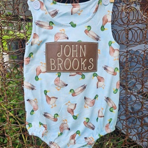 May include: A light blue baby onesie with a brown patch that says "JOHN BROOKS" and a pattern of brown ducks with green heads.