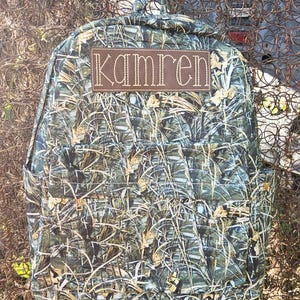 May include: A camouflage backpack with a brown leather patch that says "Kamren" on it.