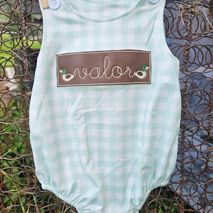 May include: A light green and white gingham baby onesie with a brown patch that says "valor" and has two embroidered ducks on either side.