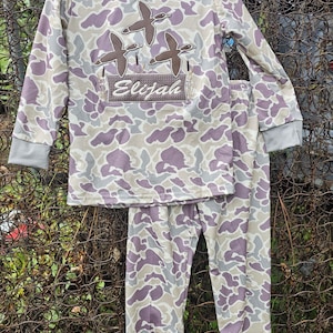 May include: A two-piece pajama set for children featuring a camouflage print in shades of purple and beige. The long-sleeved shirt has a brown applique of three ducks and the name "Elijah" in a brown banner. The pants are long and have a matching camouflage print.