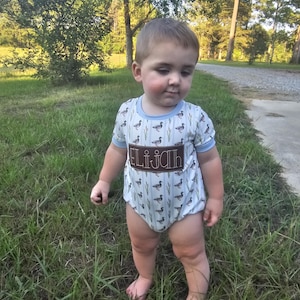 May include: A light blue baby onesie with a duck and cattail print. The onesie has a brown patch with the name "Elijah" embroidered on it.