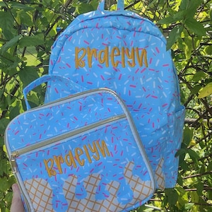 May include: A light blue backpack and matching lunch bag set. Both items feature a sprinkle pattern and the name "Braelyn" embroidered in gold. The lunch bag has a waffle cone design at the bottom.