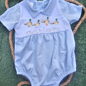 May include: A light blue baby onesie with a white collar and embroidered ducks. The onesie has the name "Adrian" embroidered on the front.