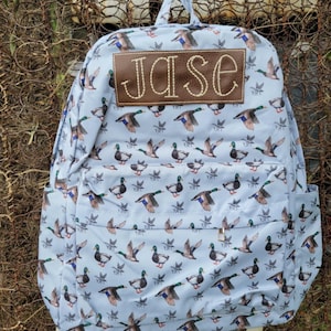 May include: A light blue backpack with a brown leather patch that says "Jase" in white stitching. The backpack has a pattern of flying ducks all over it.