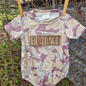 May include: A pink and brown camouflage baby onesie with the name "Branson" embroidered on the front.
