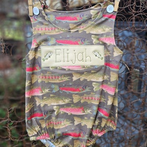 May include: A brown and green baby onesie with a pattern of trout fish. The onesie has a white button on each shoulder strap and a white embroidered name tag that says "Elijah" with two small fish on either side of the name.