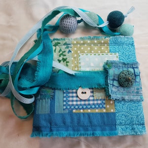 May include: A small patchwork bag made of blue and green fabric with a frayed edge. The bag has a button closure and is decorated with a blue and green button. The bag is surrounded by blue and green ribbon.
