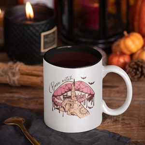 May include: White coffee mug with a black rim. The mug has a pink and black illustration of a mouth with a spiderweb and a hand with a finger over the mouth. The text "Basic witch" is written above the mouth.