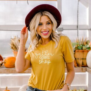 May include: A woman wearing a mustard yellow t-shirt with the text "gather gobble shop" in white. She is wearing a burgundy fedora and smiling.