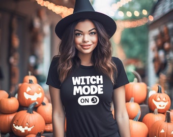 May include: A black t-shirt with the text "WITCH MODE ON" in white, worn with a black witch hat. The background features carved pumpkins and string lights, creating a Halloween theme.