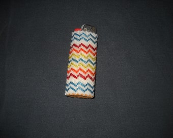 May include: A white, beaded lighter cover with a chevron pattern in red, yellow, blue, and white.