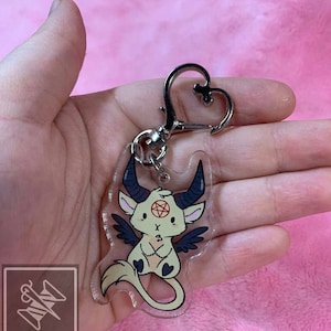 May include: A clear acrylic keychain featuring a cartoon demon with black wings, horns, and a pentagram on its forehead. The demon is white with black eyes and a pink nose. The keychain has a silver heart-shaped clasp.