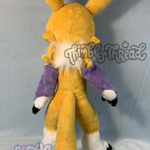 May include: A plush fox-like creature with yellow fur, white accents, and purple sleeves. The plush has large, upright ears and a fluffy white chest. The tail is long and bushy. The Thimble Thread logo is visible.