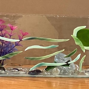 May include: A glass terrarium with a green glass sun, green and white glass seaweed, and a purple and pink plastic plant. The terrarium is filled with small white and gray stones.