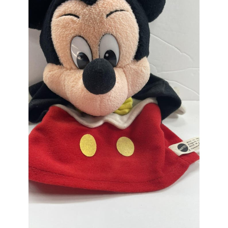 May include: A plush Mickey Mouse puppet with a black head and ears, a tan face, and large black eyes. The puppet wears a red outfit with two yellow circular buttons and a white collar. A yellow bow tie is visible. The puppet is made by Mattel.