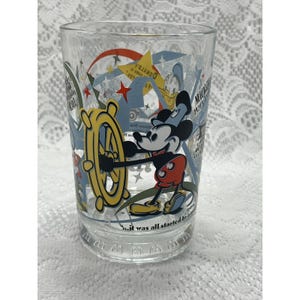 May include: A clear glass tumbler featuring a colorful graphic of Mickey Mouse at a ship's wheel. The design includes stars, geometric shapes, and text that reads "It all was started by a mouse" along with "Mickey 1928" and "Donald".