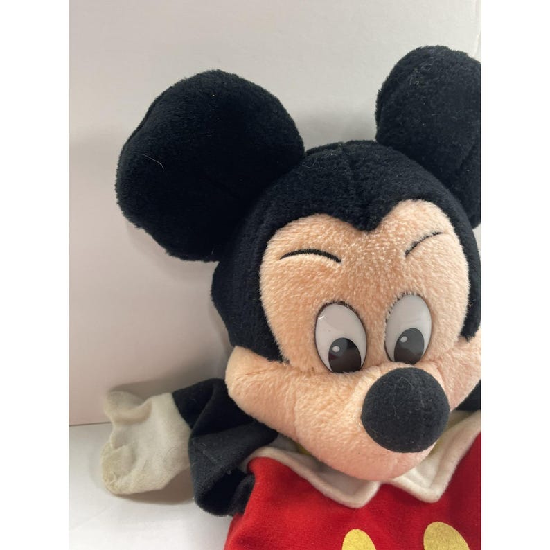 May include: A plush Mickey Mouse doll with black ears, a black head, and a tan face. The doll is wearing a red shirt with yellow buttons and white gloves. The doll's eyes are large and white with black pupils.