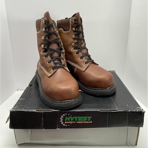 May include: A pair of brown leather work boots with black laces, sitting on a black and white box. The boots have a padded collar and a sturdy sole. The box has the text "HYTEST SAFETY FOOTWEAR".