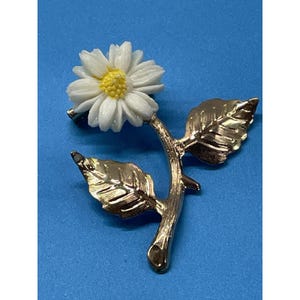May include: A gold-toned metal brooch shaped like a flower stem with two textured leaves. The flower head features white petals surrounding a yellow center, creating a detailed botanical design that is secured with a pin on the back side.