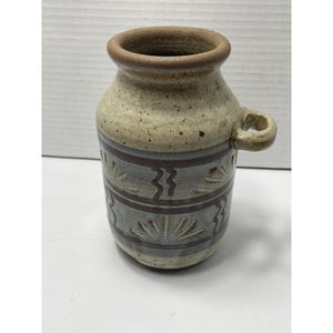 May include: A ceramic pitcher with a speckled cream-colored upper section and a grey lower section featuring dark brown horizontal bands. The body is decorated with embossed sunburst and wavy line patterns and includes a single side handle.