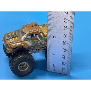 May include: A brown and gold monster truck toy with large black tires. The truck has a detailed design with a cartoon graphic on the side. The ruler shows the truck is approximately 3 inches long.
