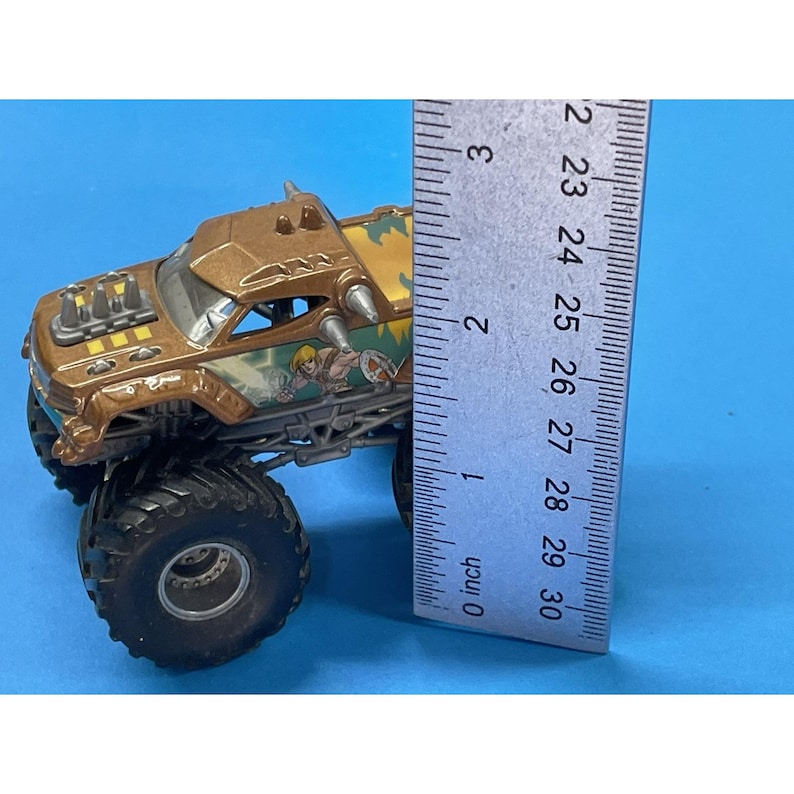 May include: A brown and gold monster truck toy with large black tires. The truck has a detailed design with a cartoon graphic on the side. The ruler shows the truck is approximately 3 inches long.