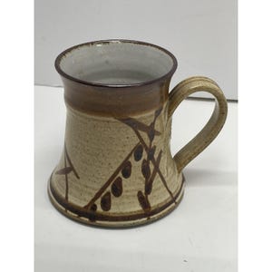 May include: A handmade ceramic mug with a beige speckled finish. The mug features a brown rim and handle, with a decorative pattern of brown lines and dots. The mug has a flared shape and is suitable for hot or cold beverages.